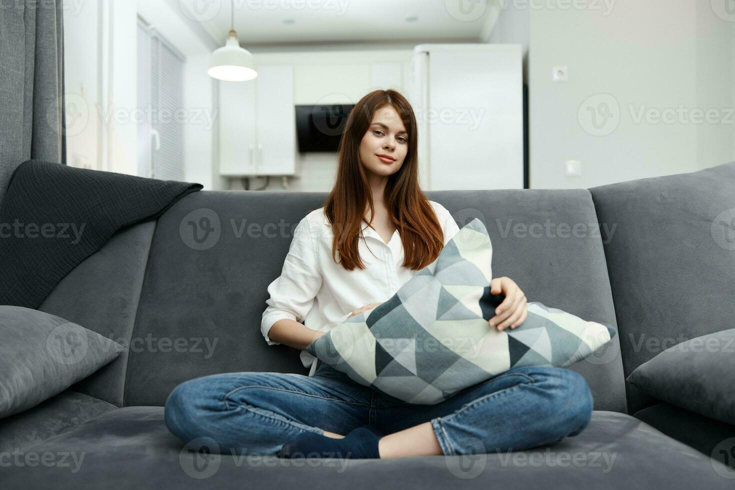 femme séance sur le canapé dans sa gratuit temps avec une oreiller dans sa mains du repos appartement intérieur photo