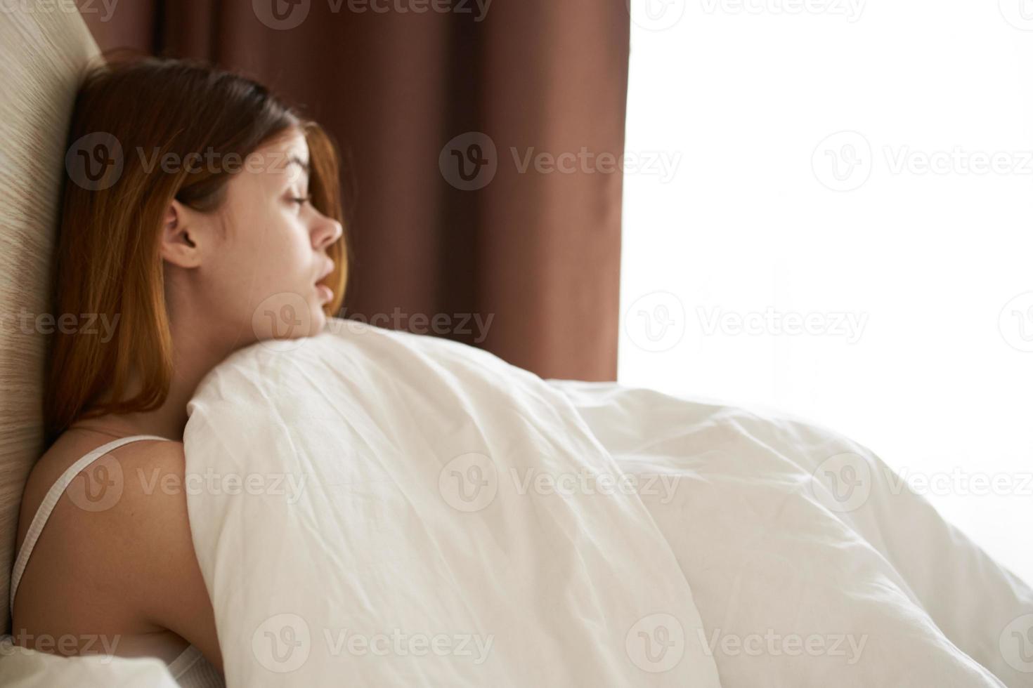 une jolie femme dans une nuit costume est mensonge dans lit en dessous de une couverture près le fenêtre sur une fin de semaine vacances photo