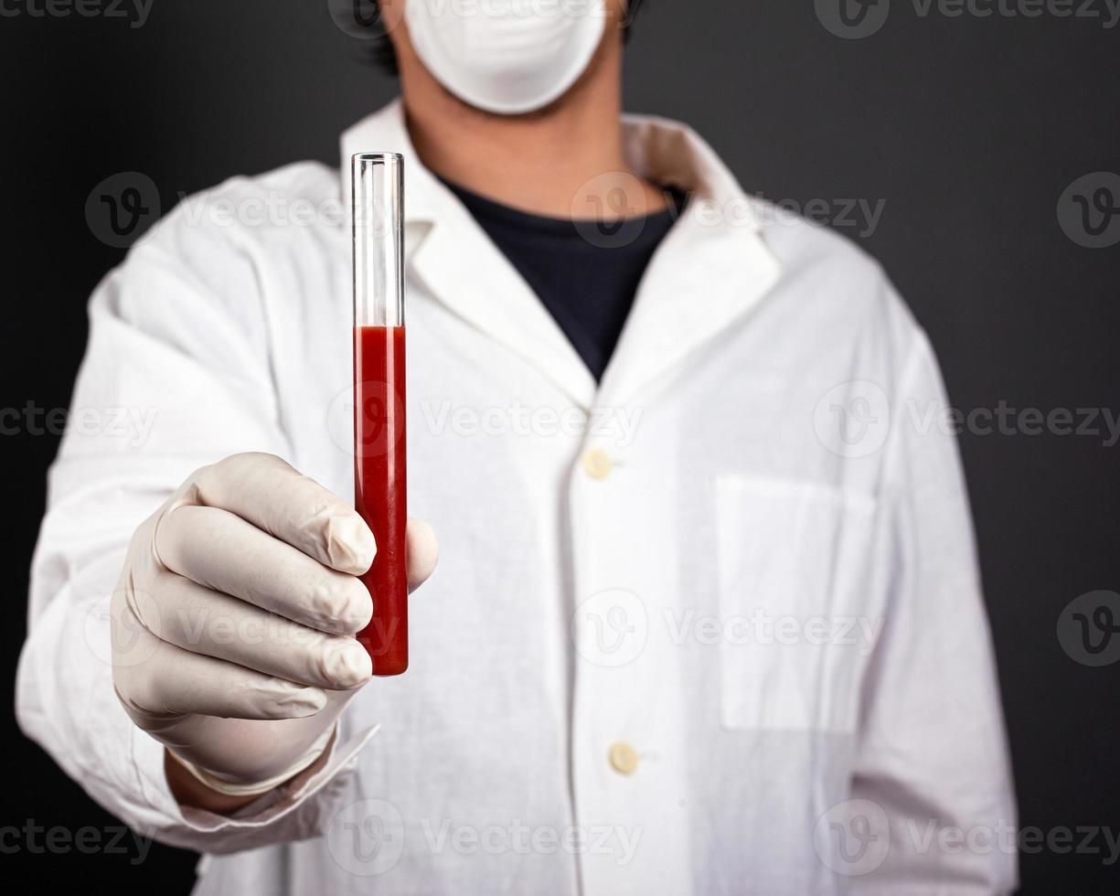 médecin en blouse blanche avec des gants jetables et un masque médical tenant un tube à essai dans sa main avec du sang infectieux. photo