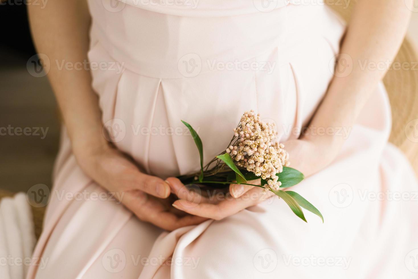 une Enceinte femme détient une petit bouquet de fleurs dans sa mains. une Jeune femme dans une délicat rose maternité robe est attendre pour le naissance de une enfant. grossesse, maternité photo