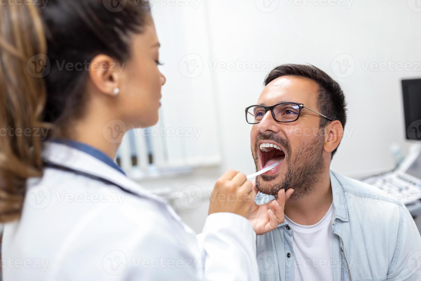 une Jeune homme est assis sur un examen table à travers de le sien médecin. le médecin atteint vers l'avant avec une langue dépresseur comme le homme regards en haut et des bâtons en dehors le sien langue. photo