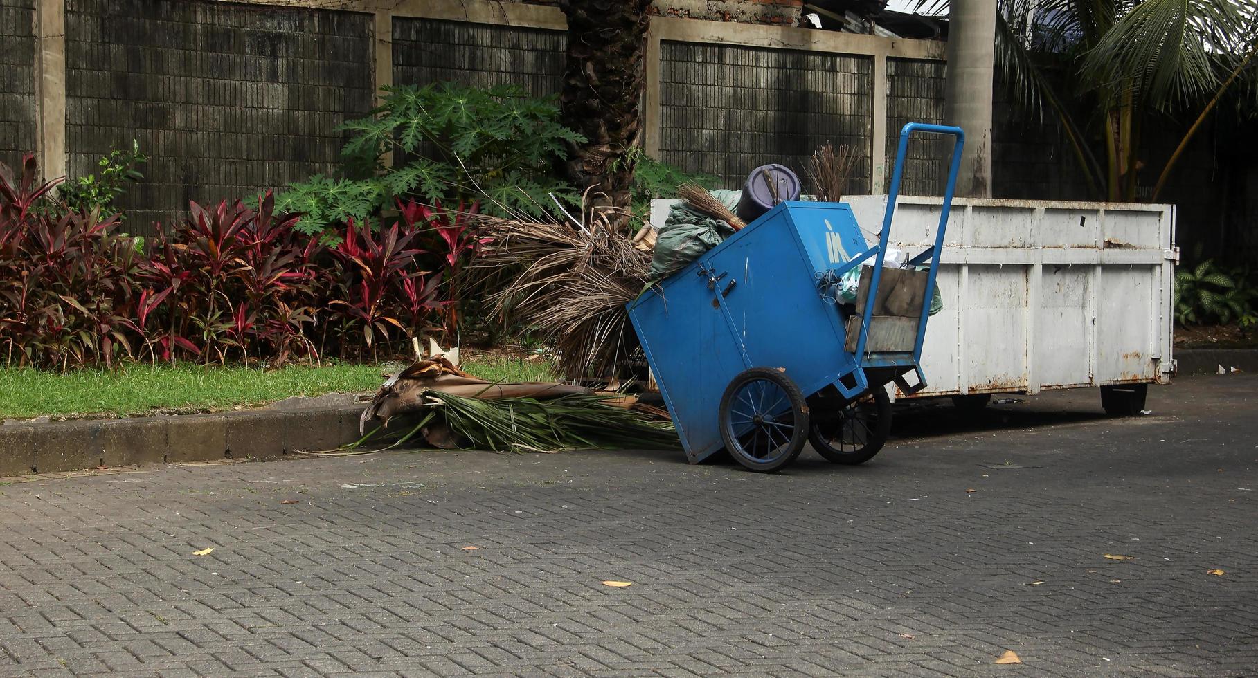 un bleu tirer poubelle Chariot avec roues. gerobak sampah Biru la photographie objet Extérieur isolé sur minuscule jardin et brique sol sol Contexte. photo