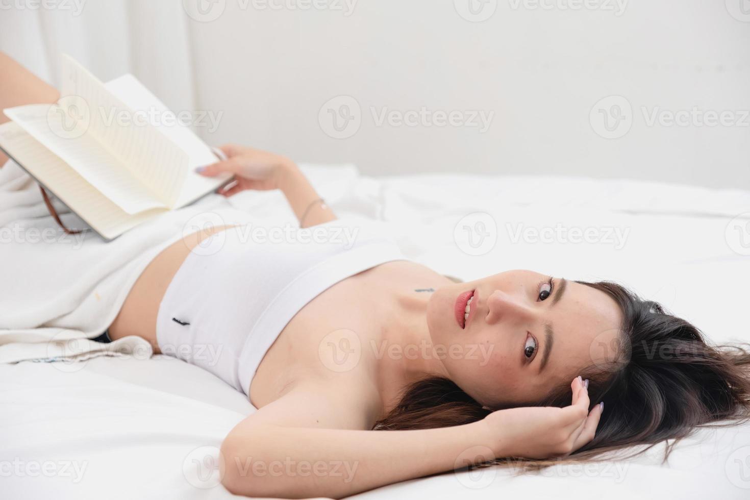 portrait de blanc asiatique femme avec tatouage repos dans lit à du repos et se détendre à l'intérieur blanc chambre sur sa vacances, vacances Matin concept. photo