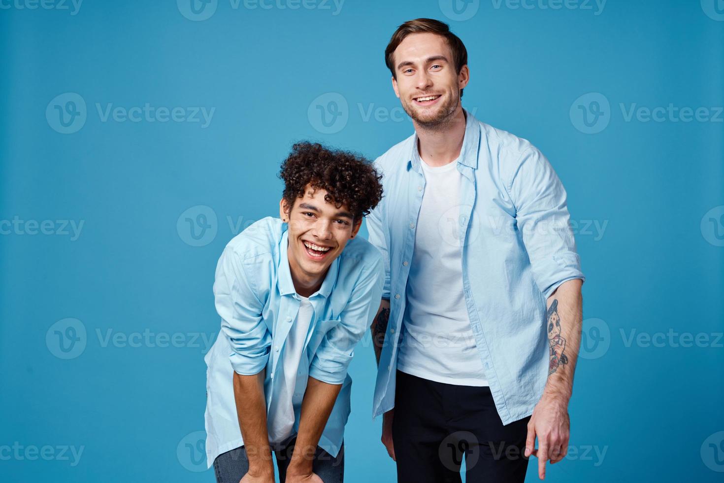 aux cheveux bouclés gars dans une bleu chemise penché vers l'avant et une blond homme sur une bleu Contexte copains photo