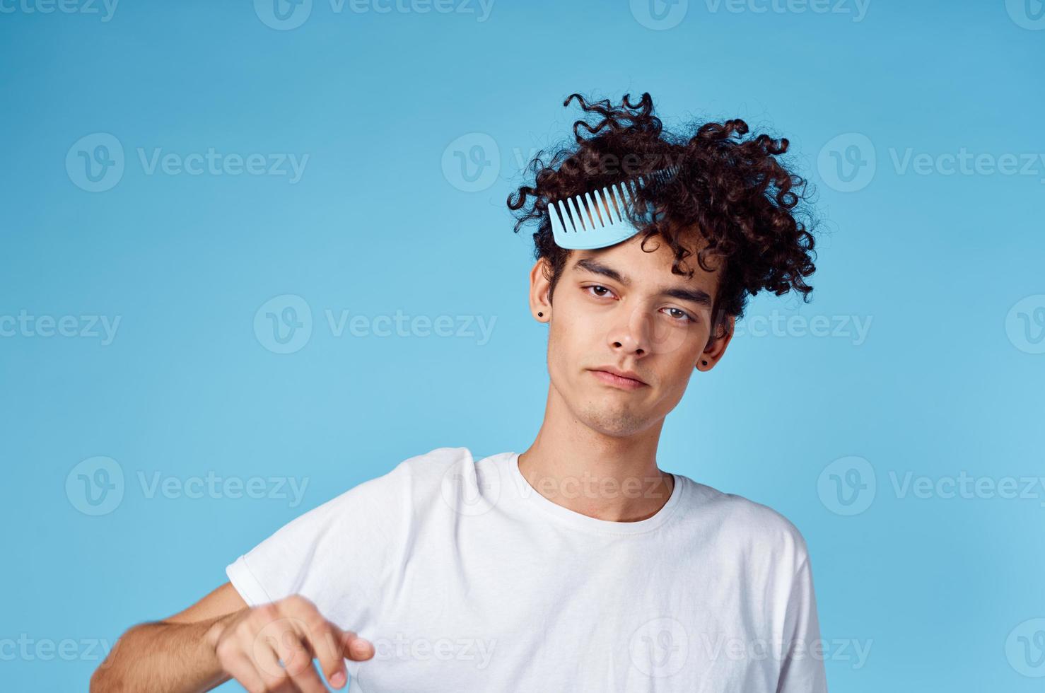 brunette gars avec frisé cheveux sur une bleu Contexte et une peigne dans le sien main photo