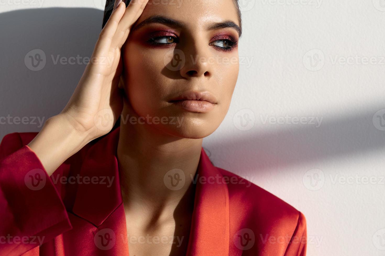 brunette avec soir maquillage et dans une rouge veste tenir sa main près sa tête photo