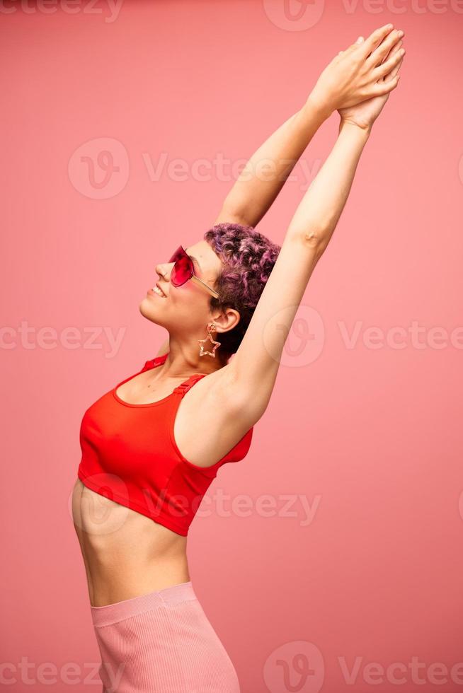 mode portrait de une femme avec une court la Coupe de cheveux dans coloré des lunettes de soleil avec inhabituel accessoires avec des boucles d'oreilles souriant sur une rose brillant Contexte avec une aptitude corps dansant photo