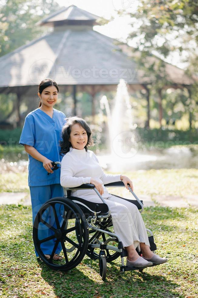 personnes âgées asiatique Sénior femme sur fauteuil roulant avec asiatique prudent soignant. allaitement Accueil hôpital jardin concept. dans lumière du soleil photo