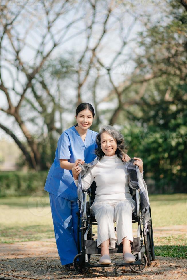 asiatique prudent soignant ou infirmière et le content patient dans une fauteuil roulant sont en marchant dans le jardin. à Aidez-moi et encourager et du repos votre esprit avec vert la nature. utilisation une stéthoscope à Ecoutez à votre photo