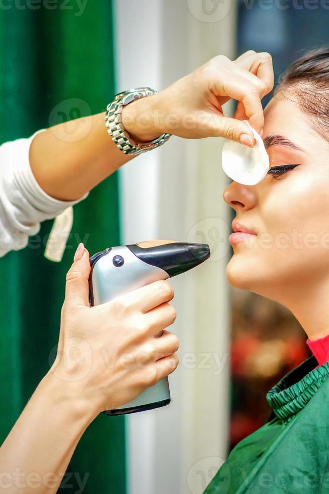 côté vue de une maquillage artiste en utilisant aérographe fabrication un aérographe maquillage fondation sur une femelle visage dans une beauté salon. photo