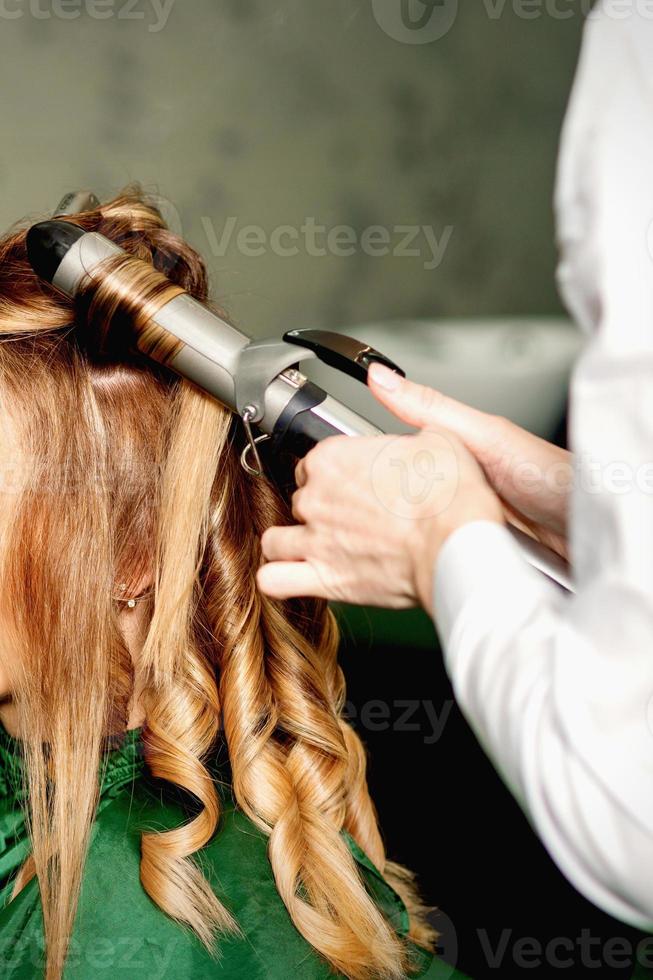 coiffeur en utilisant curling pinces boucles longue marron cheveux sur le Jeune caucasien fille dans une beauté salon. photo