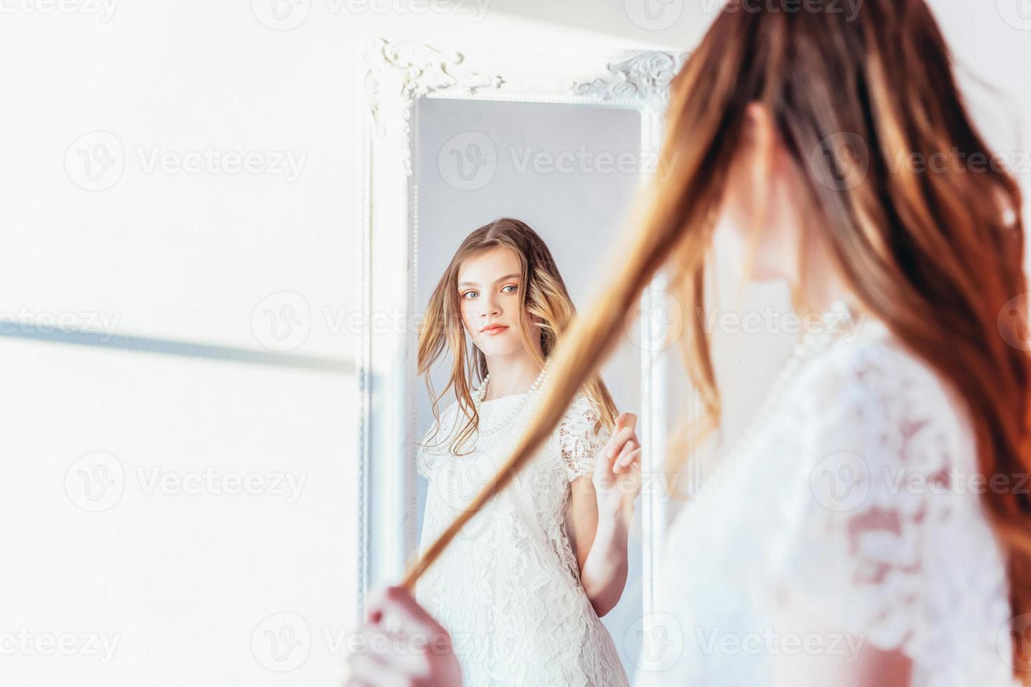 beauté maquillage Matin rutine l'amour toi même concept. Jeune adolescent fille à la recherche à réflexion dans miroir. Jeune positif femme portant blanc robe posant dans brillant lumière pièce contre blanc mur. photo