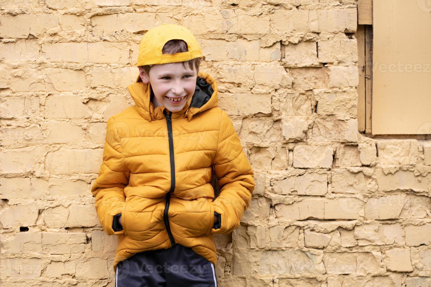 une enfant sur le rue. une content garçon est permanent contre une brique mur dans Jaune vêtements et une base-ball casquette. photo