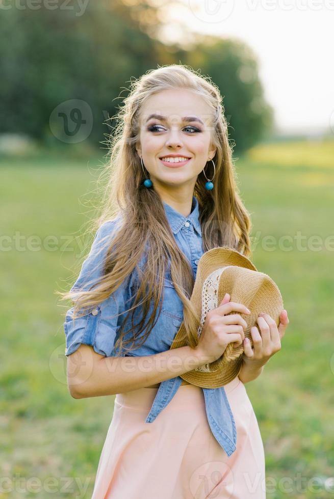 souriant Jeune femme détient une chapeau dans sa mains tandis que en marchant dans le été en plein air à l'extérieur le ville sur vacances photo