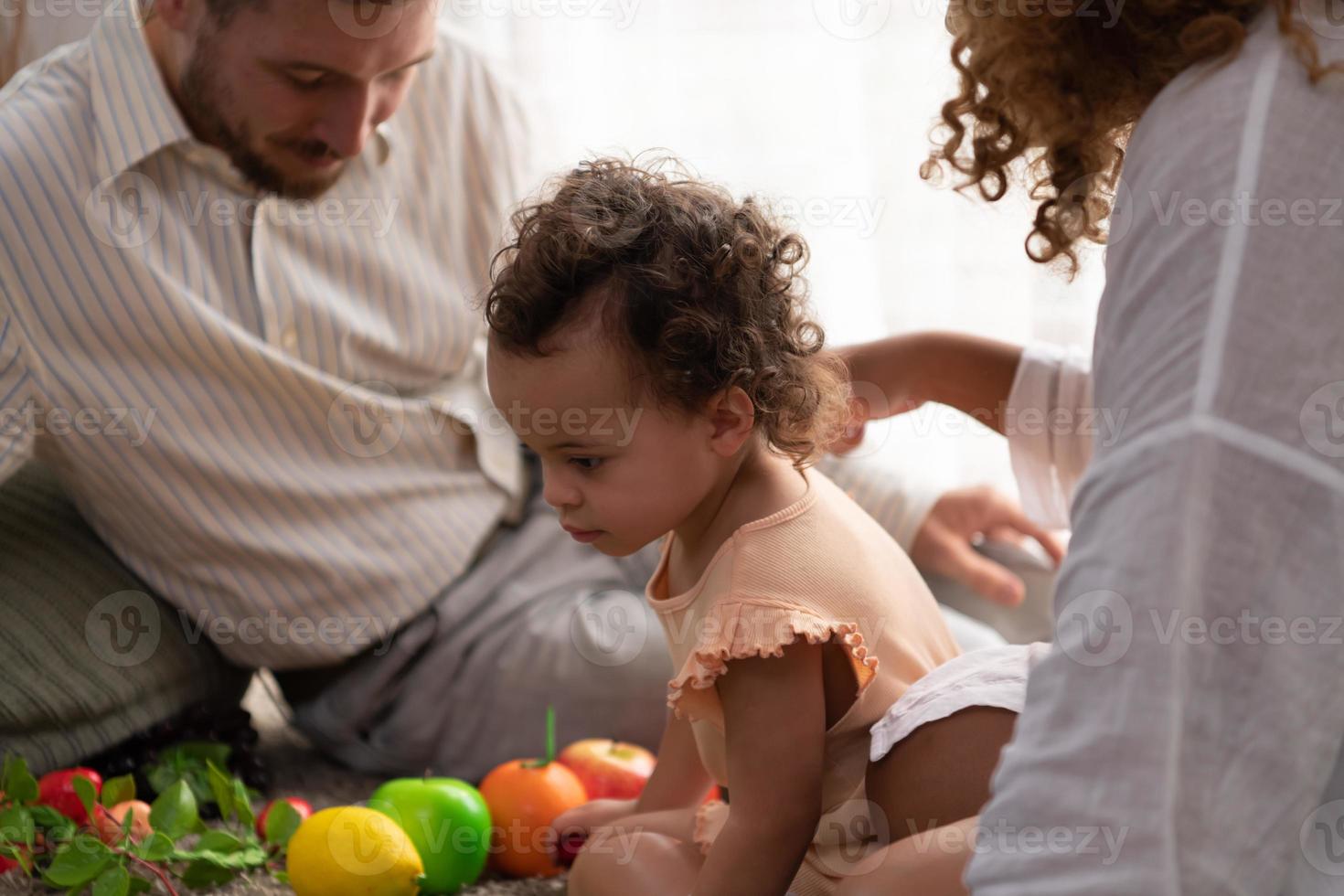 Parents et les enfants se détendre dans le vivant pièce de le maison. regarder bébé Heureusement jouer avec le sien préféré jouet. photo