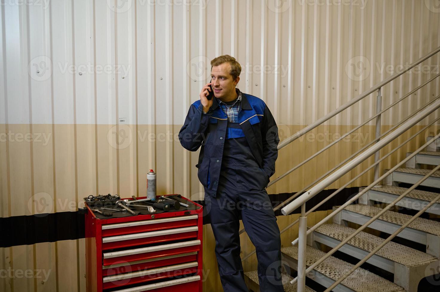 portrait de une Beau caucasien homme, technicien, garage mécanicien parlant sur mobile téléphone permanent près une boîte avec travail outils pour inspecter et réparation automobiles et voiture un service station photo