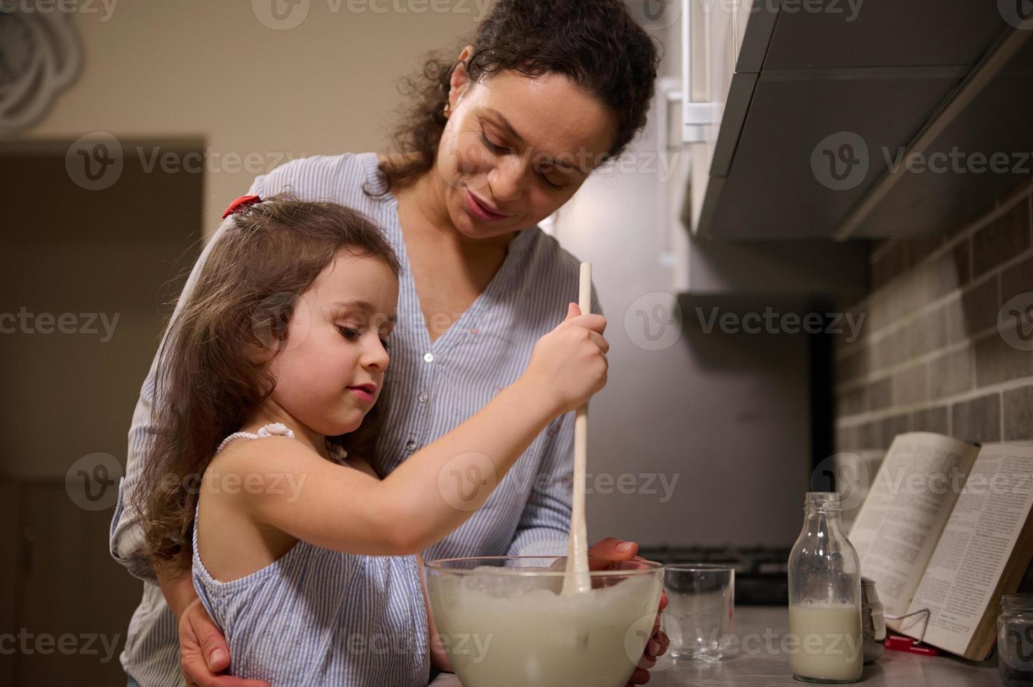 adorable peu fille ayant amusant, concentré sur mélange Ingrédients et pétrissage pâte dans une verre bol tandis que sa aimant content mère enseignement sa à cuisinier crêpe pâte. rétrécir Mardi concept photo