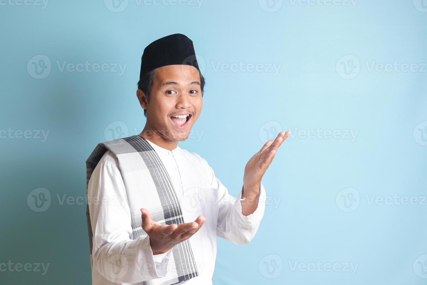 portrait de asiatique musulman homme dans blanc koko chemise avec calotte tirant mains vers caméra, attrayant Quelqu'un à viens à l'intérieur, accueillant geste. isolé image sur bleu Contexte photo