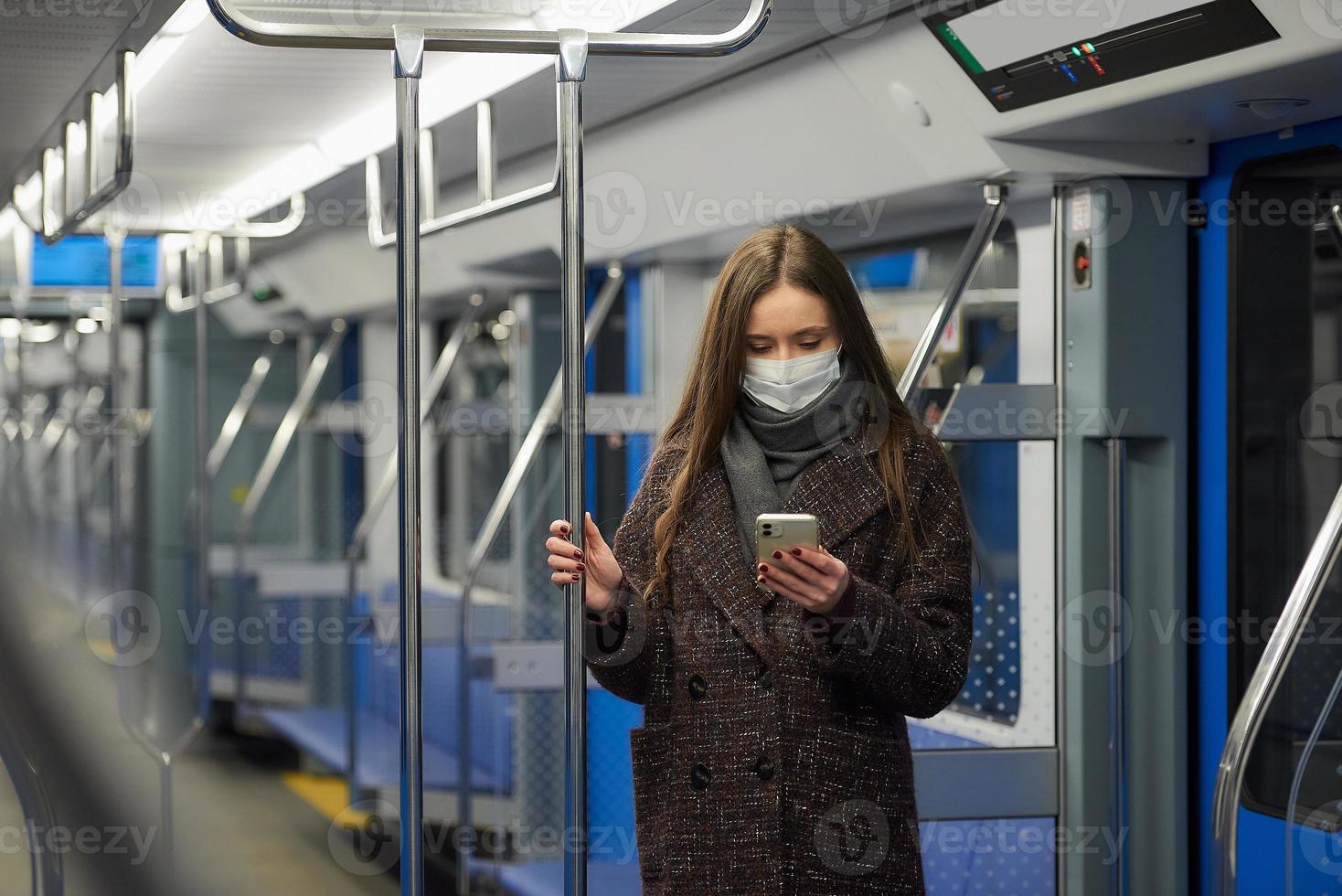 Une femme dans un masque facial est debout et à l'aide d'un smartphone dans une voiture de métro moderne photo