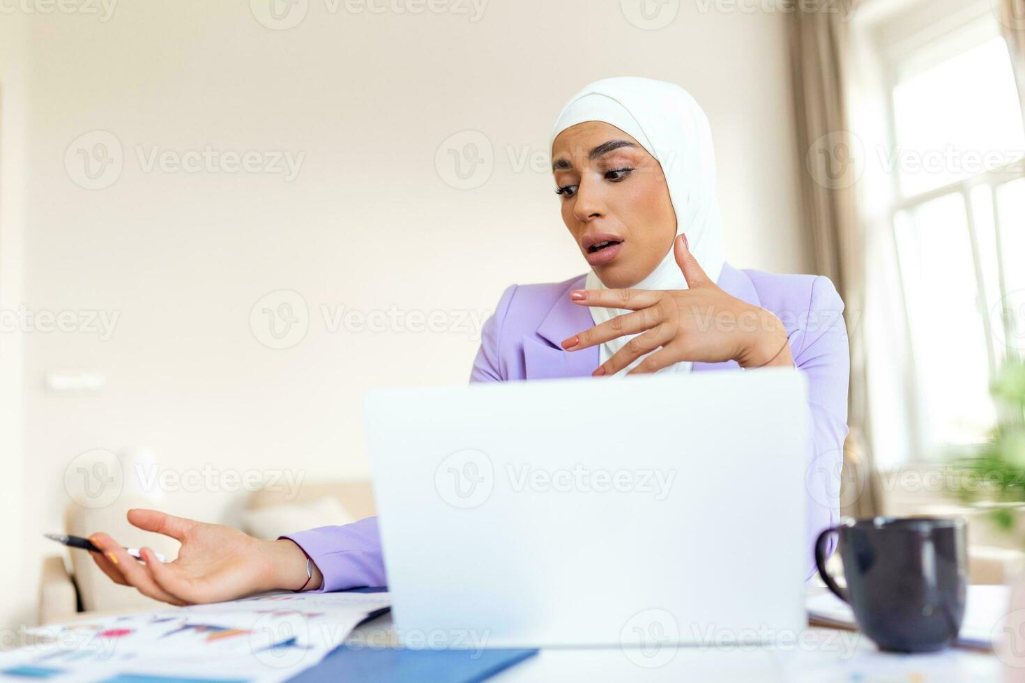 femme musulmane travaillant avec ordinateur. jeune femme d'affaires arabe assise à son bureau à la maison, travaillant sur un ordinateur portable et buvant du café ou du thé. femme musulmane travaillant à la maison et utilisant un ordinateur. photo