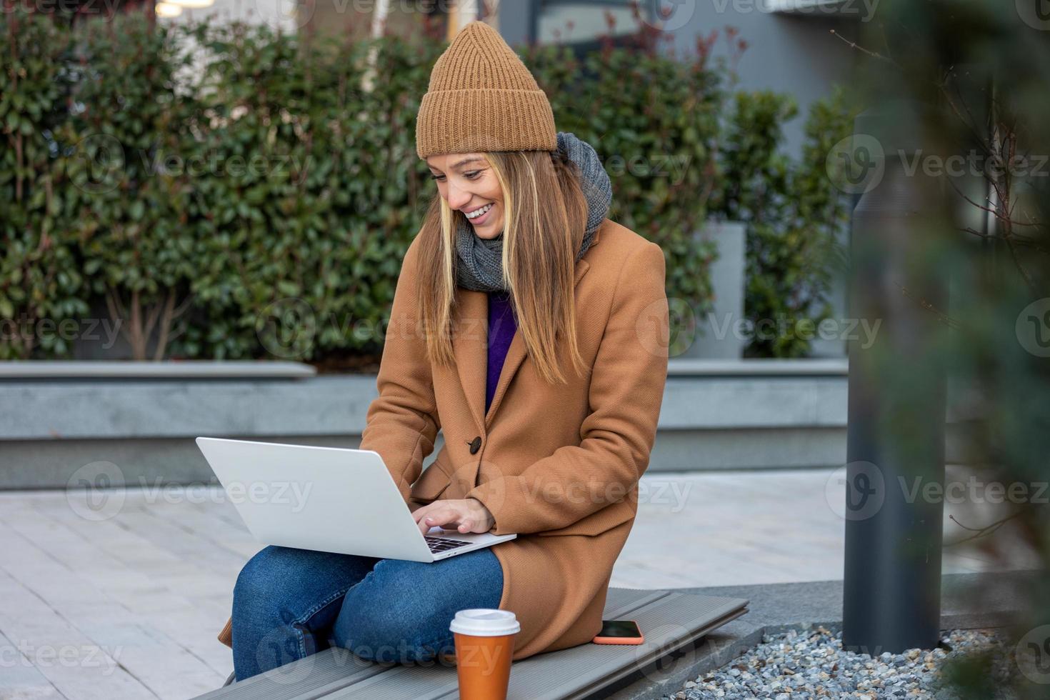 attrayant caucasien fille travail sur une ville rue sur une banc elle détient une portable dans sa mains, derrière sa une moderne bâtiment, Bureau ouvrier, pigiste. photo