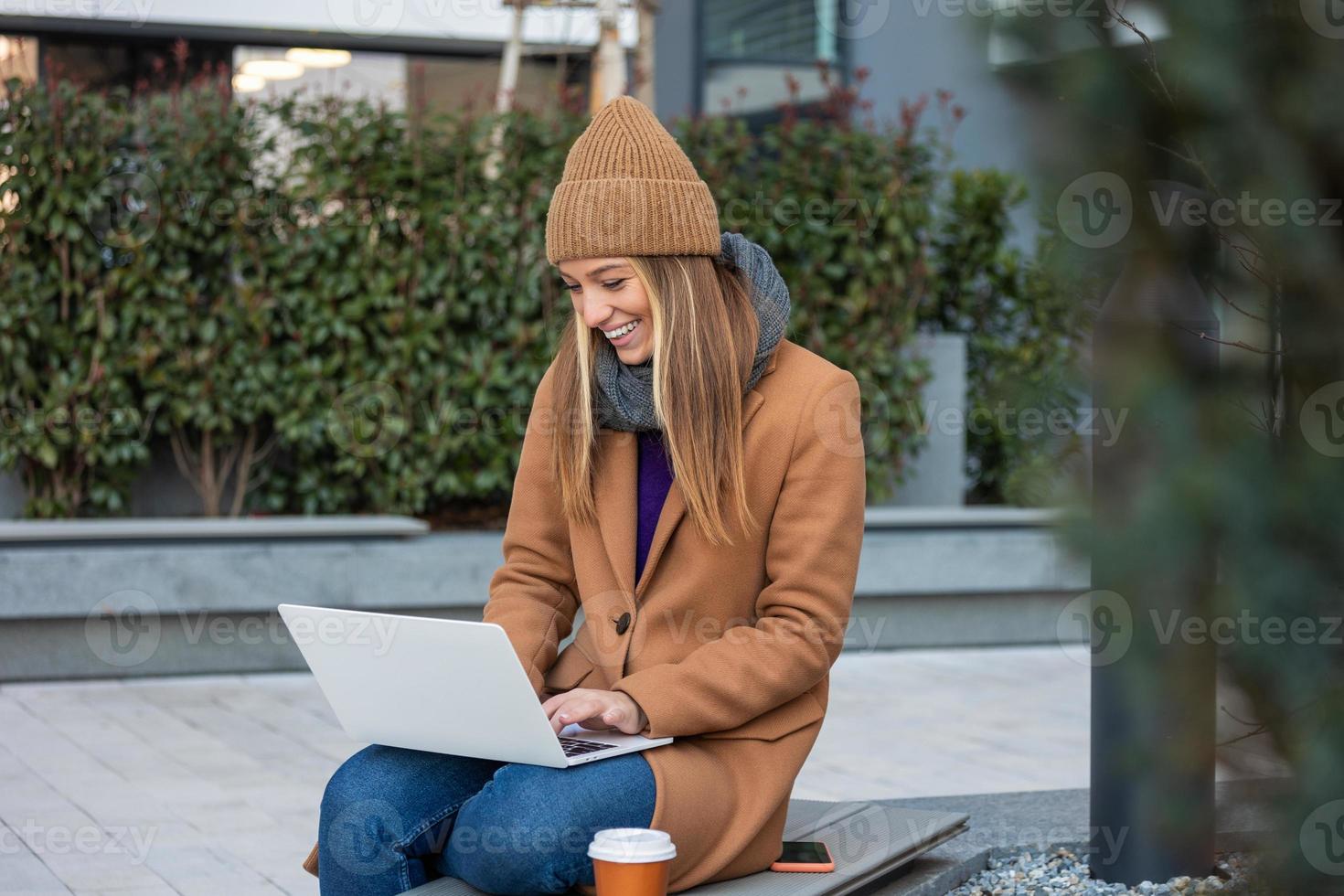 attrayant caucasien fille travail sur une ville rue sur une banc elle détient une portable dans sa mains, derrière sa une moderne bâtiment, Bureau ouvrier, pigiste. photo