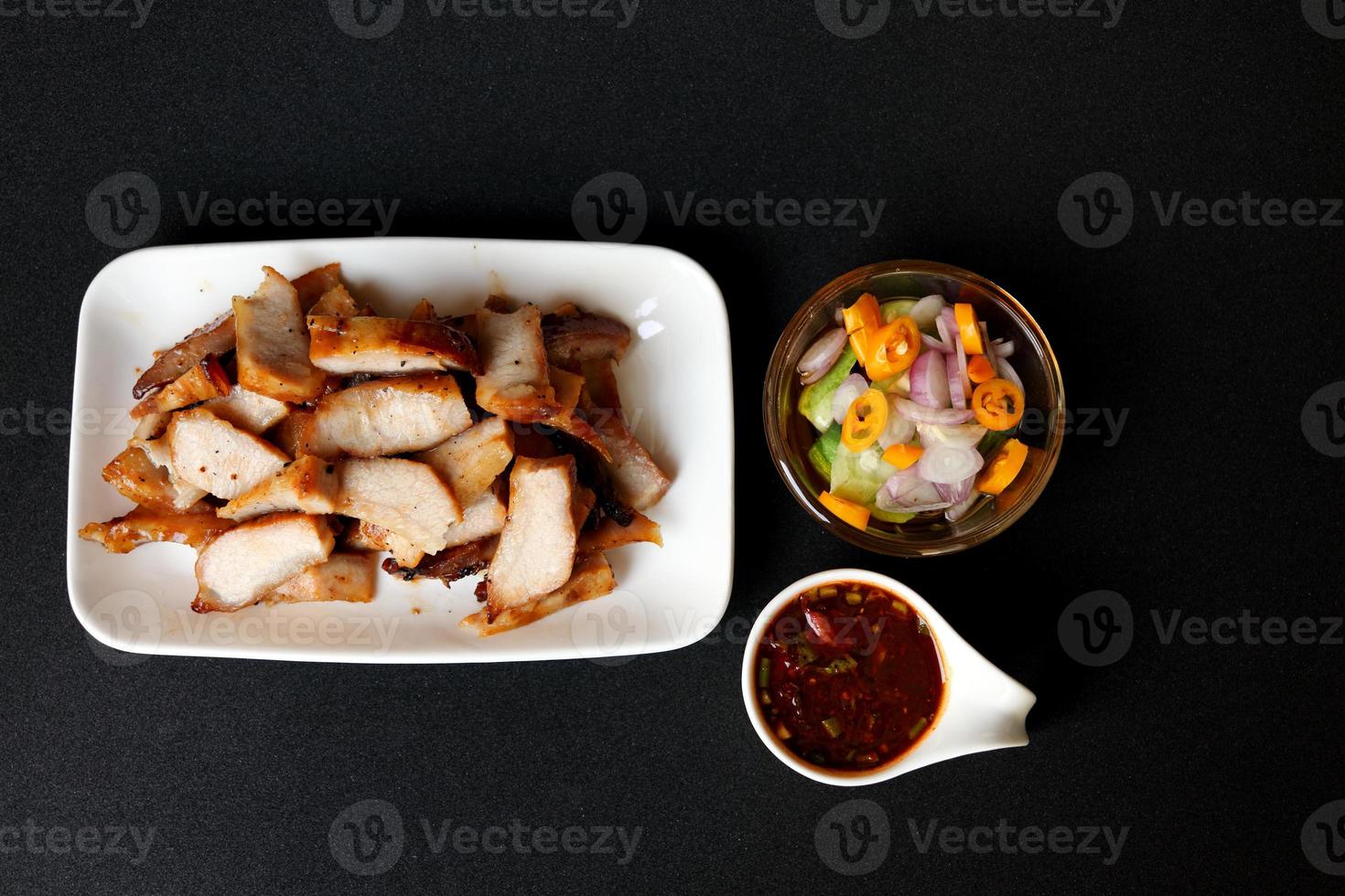 grillé porc épaule tranche dans blanc rectangle plat avec chaud et acide thaïlandais style sauce dans blanc tasse et légume tranche dans le vinaigre verre tasse sur noir Contexte. photo