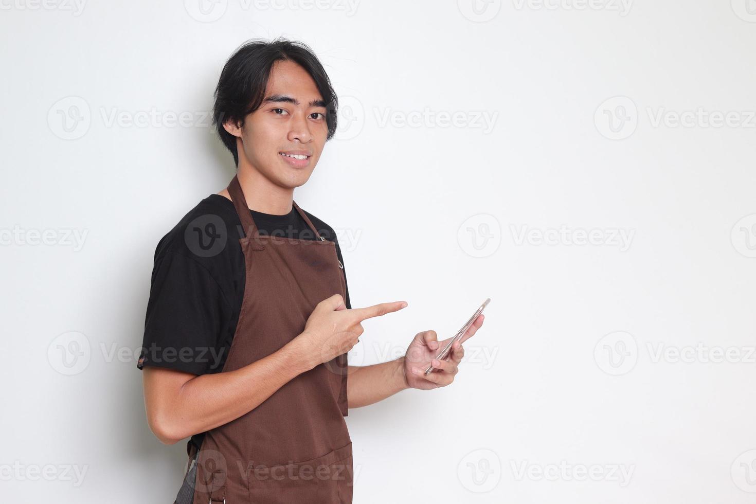 portrait de attrayant asiatique barista homme dans marron tablier en portant et montrer du doigt mobile téléphone. isolé image sur blanc Contexte photo