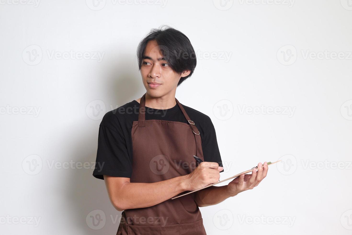 portrait de attrayant asiatique barista homme dans marron tablier prise commande, l'écriture sur menu livre liste. isolé image sur blanc Contexte photo