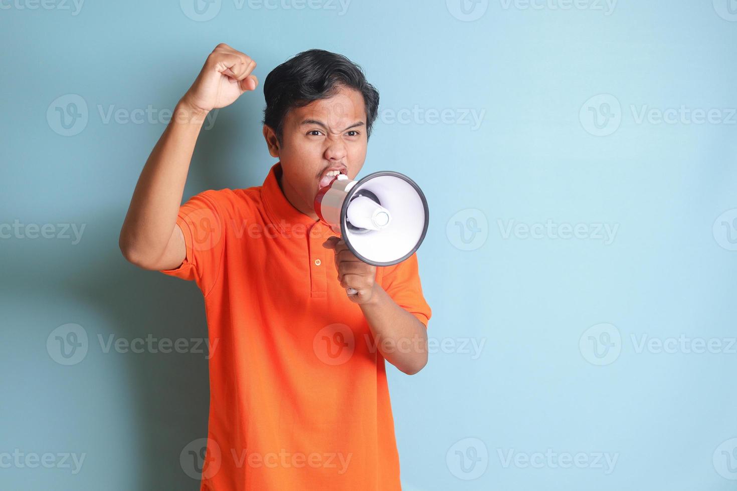 portrait de attrayant asiatique homme dans Orange chemise Parlant Plus fort en utilisant mégaphone, promouvoir produit. La publicité concept. isolé image sur bleu Contexte photo