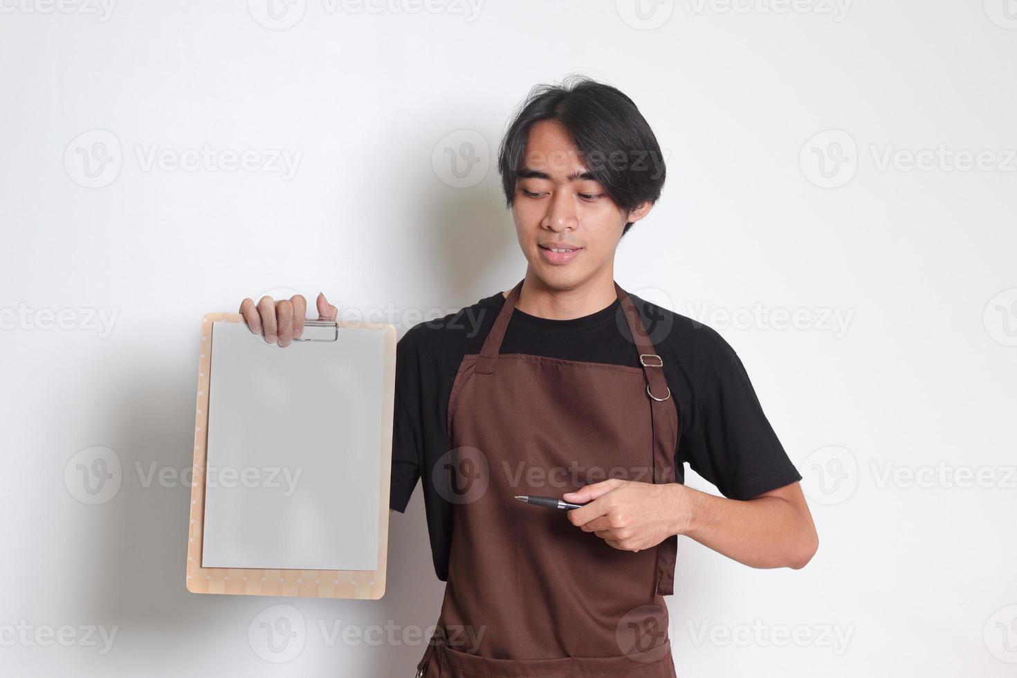 portrait de attrayant asiatique barista homme dans marron tablier montrant Vide papier de menu sur presse-papiers. isolé image sur blanc Contexte photo