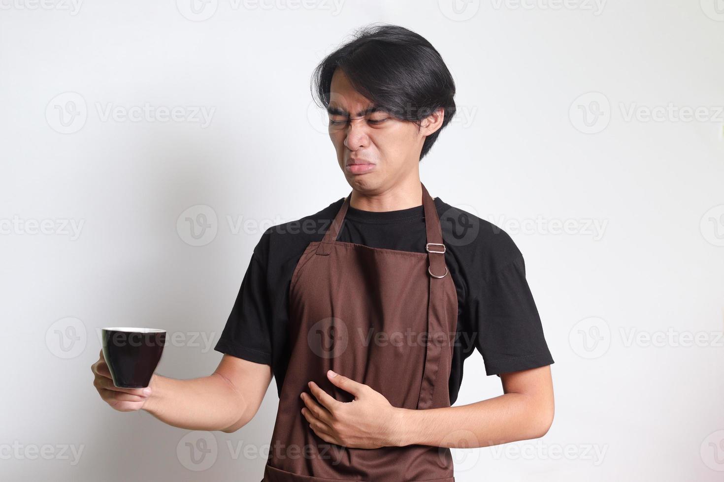 portrait de attrayant asiatique barista homme dans marron tablier Souffrance de estomac douleur en buvant une tasse de café. isolé image sur blanc Contexte photo