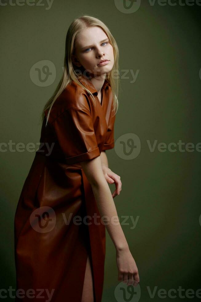 magnifique jolie confiance Jeune blond modèle dans élégant cuir marron palette robe tranchée manteau posant isolé sur olive vert studio mur Contexte. expressif femme regards à caméra. mode offre photo