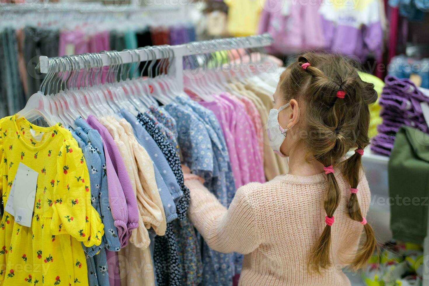 une peu fille dans une médical masque choisit une robe dans le magasin. une enfant est achats à une Vêtements centre commercial parmi le covid-19 pandémie. photo