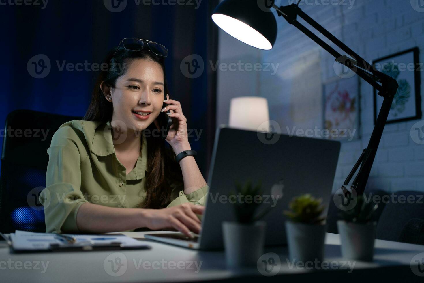 femme dans des lunettes parlant sur téléphone intelligent, travail sur bureau PC en retard à nuit à Accueil bureau, pouvez ne pas dormir, éloigné travail. photo