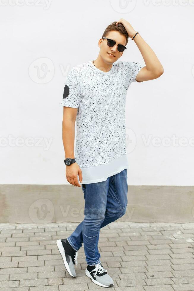 Jeune Beau homme dans une blanc T-shirt, bleu jeans et baskets permanent près une blanc mur. photo