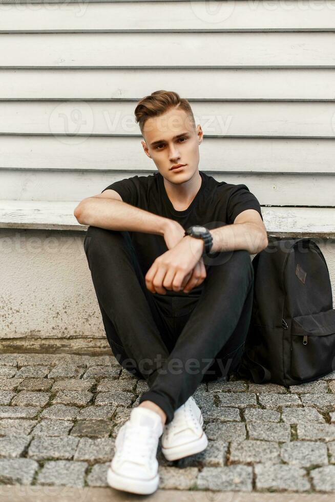 Beau branché homme dans une noir chemise, noir jeans et blanc des chaussures avec une sac à dos séance près le mur. photo