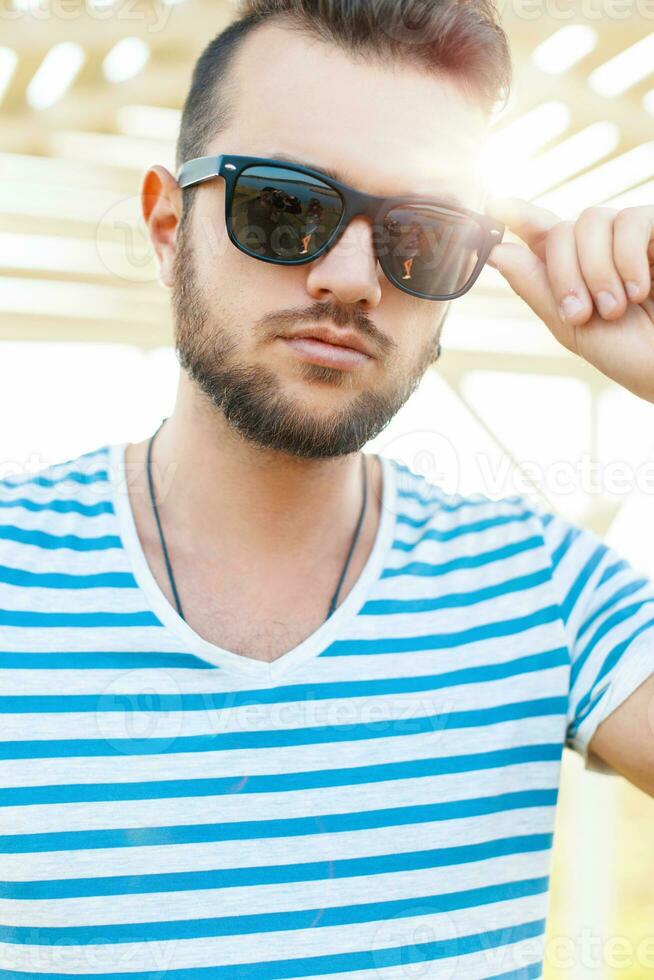 Beau branché gars avec une barbe et avec des lunettes de soleil dans le lumière du soleil sur le plage sur une été journée. photo