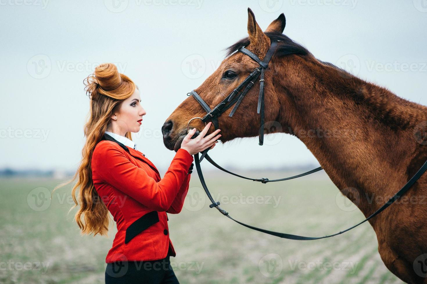 Fille jockey aux cheveux roux dans un cardigan rouge et des bottes noires avec un cheval photo