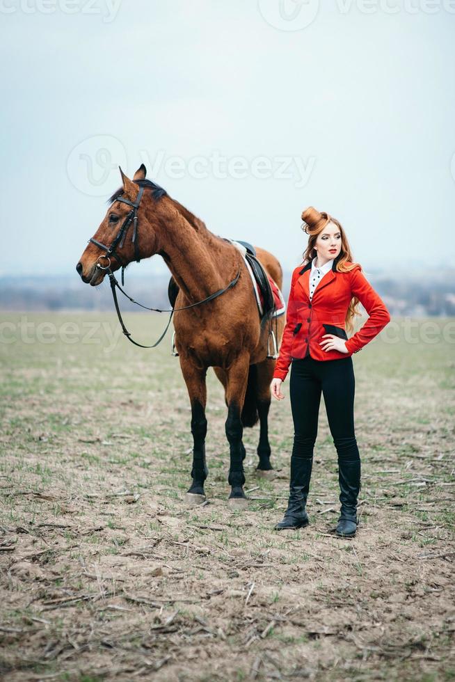 Fille jockey aux cheveux roux dans un cardigan rouge et des bottes noires avec un cheval photo