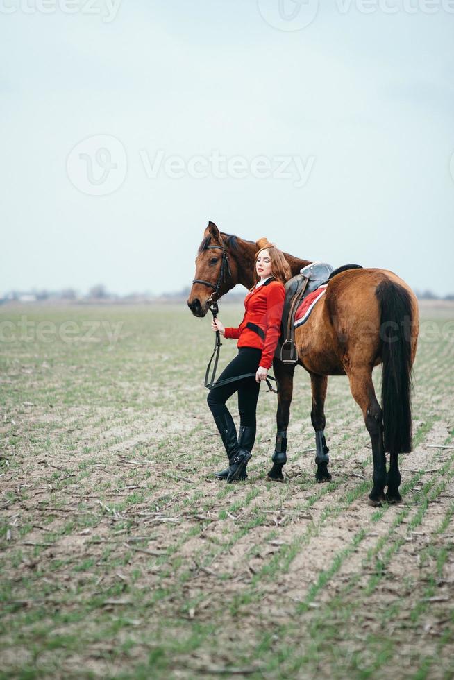 Fille jockey aux cheveux roux dans un cardigan rouge et des bottes noires avec un cheval photo