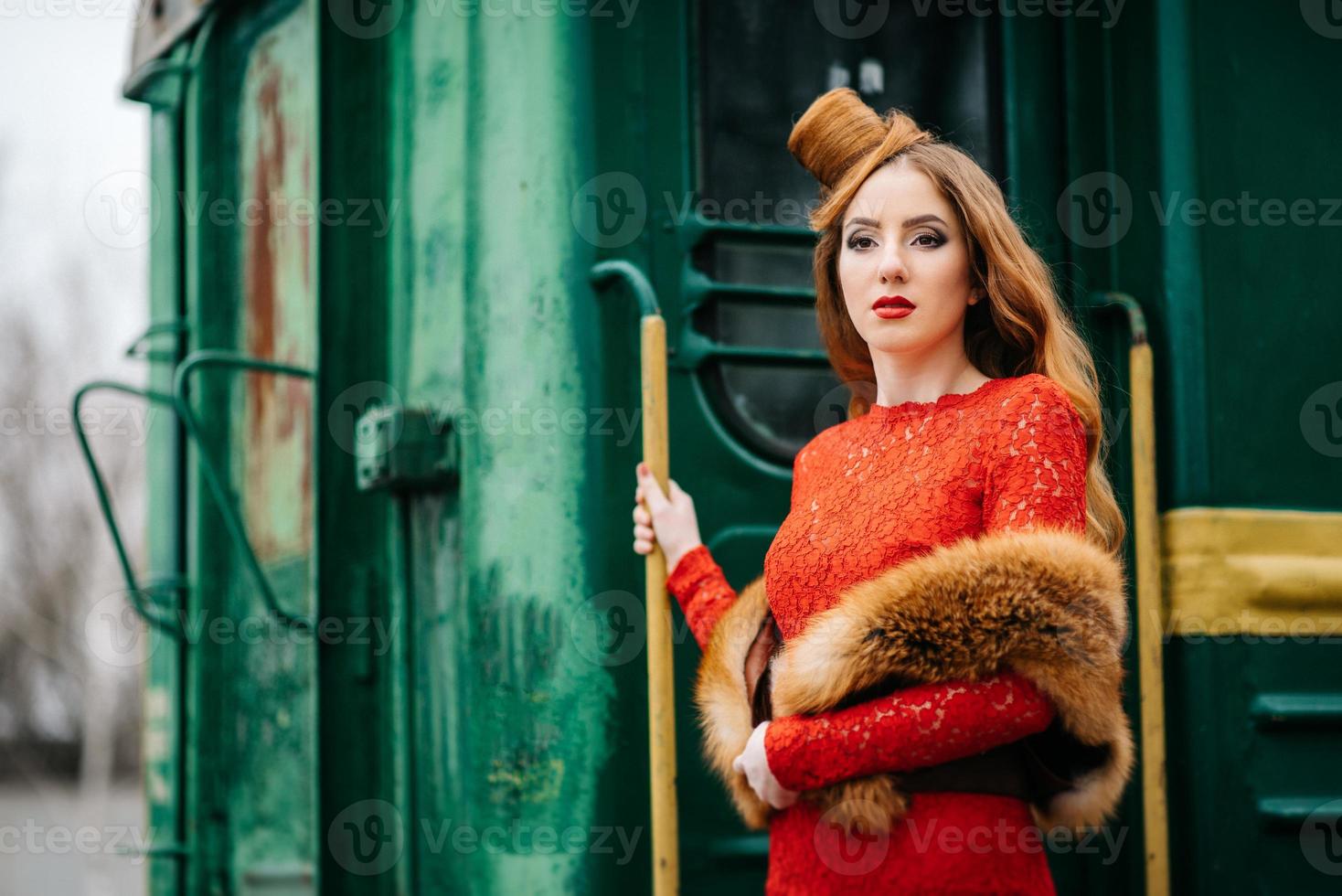 Jeune fille aux cheveux rouges dans une robe rouge vif près d'une vieille voiture de tourisme photo