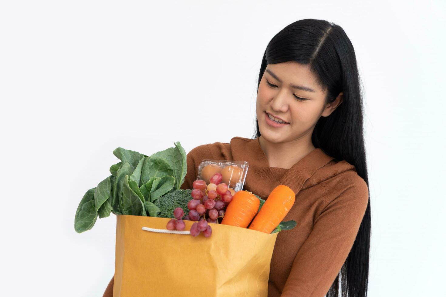 content asiatique femme est souriant et porte une achats papier sac après le courrier de le épicerie venu à livrer le sien des biens à maison. concept de supermarché livraison pour une Nouveau mode de vie photo