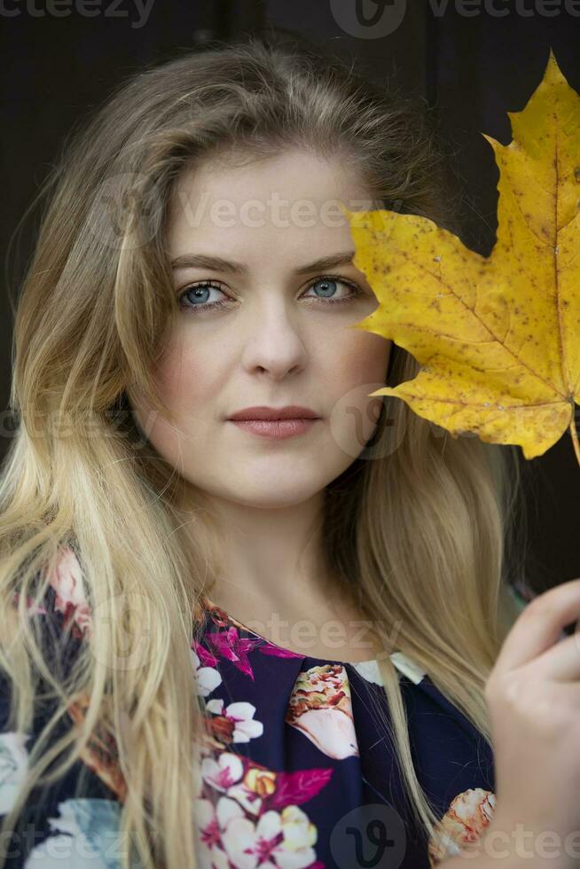 proche en haut est le visage de une magnifique fille en portant une Jaune érable feuille. aux yeux bleus blond dans l'automne. photo