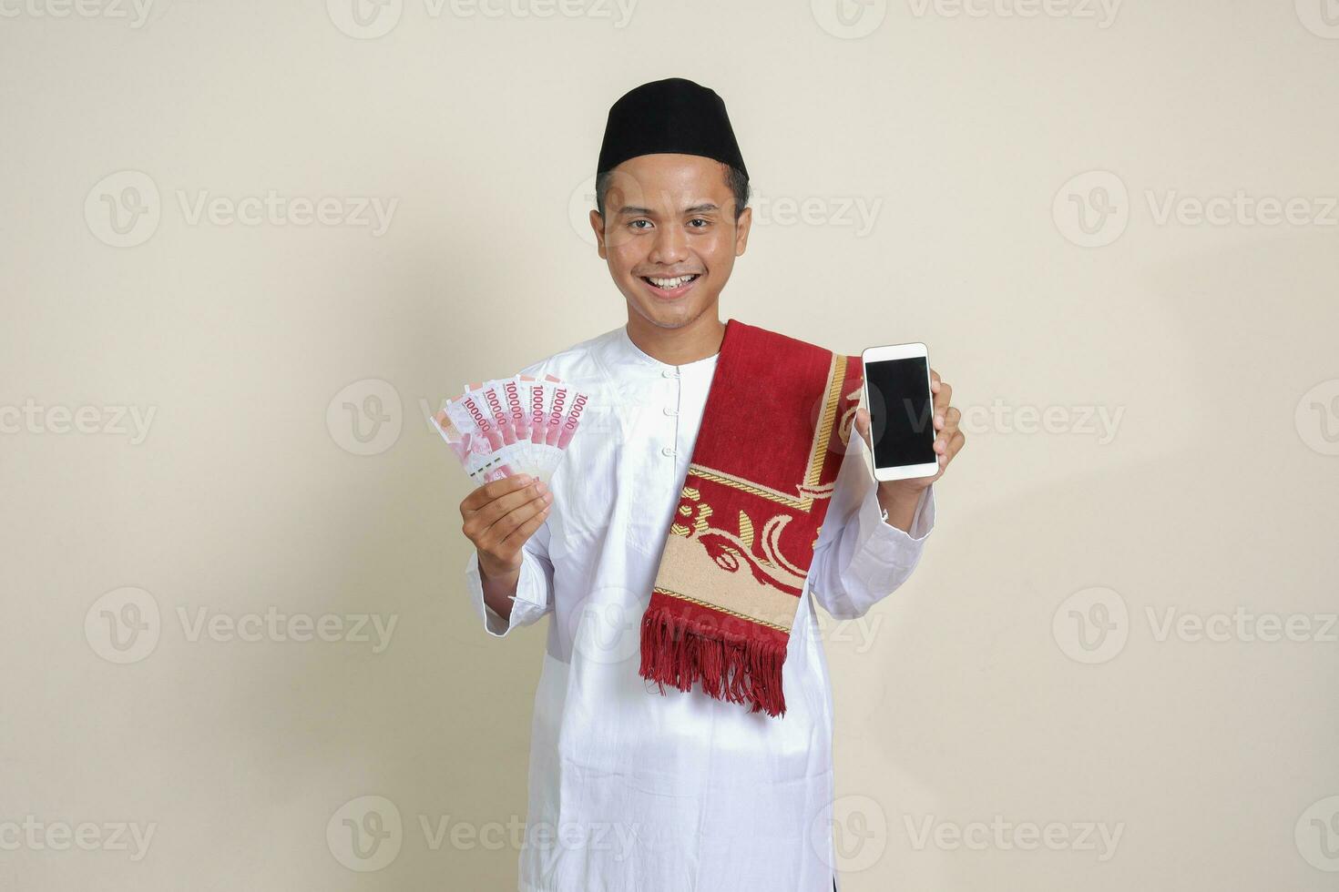 portrait de attrayant asiatique musulman homme dans blanc chemise avec calotte montrant un cent mille Rupiah tandis que montrant Vide écran mobile téléphone. financier et achats concept. isolé image sur gris photo