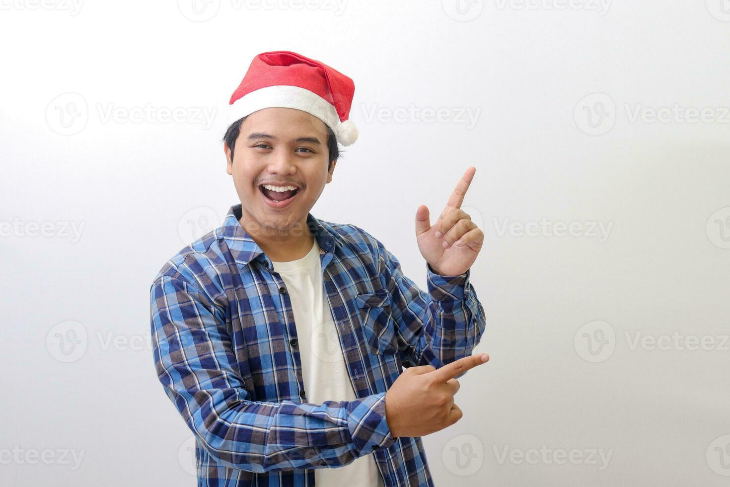 portrait de excité asiatique homme dans bleu plaid chemise portant Père Noël chapeau souriant et à la recherche à le caméra montrer du doigt avec deux mains et les doigts à le côté. isolé image sur blanc Contexte photo