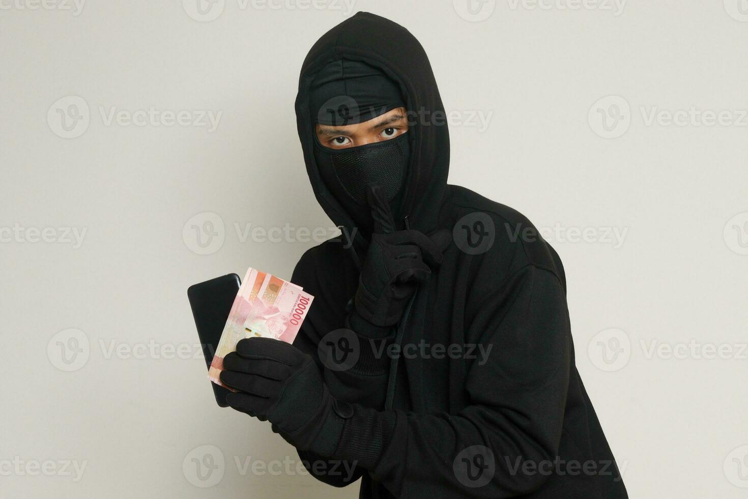 portrait de mystérieux homme portant noir sweat à capuche et masque vol un cent mille Rupiah avec succès de victime. isolé image sur gris Contexte photo
