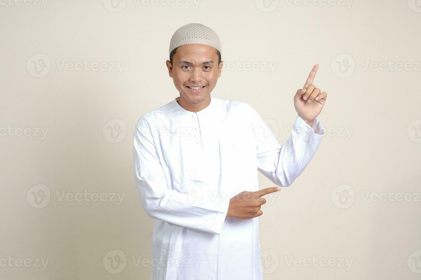 portrait de attrayant asiatique musulman homme dans blanc chemise avec calotte montrant produit et montrer du doigt avec le sien main et doigt à le côté. La publicité concept. isolé image sur gris Contexte photo