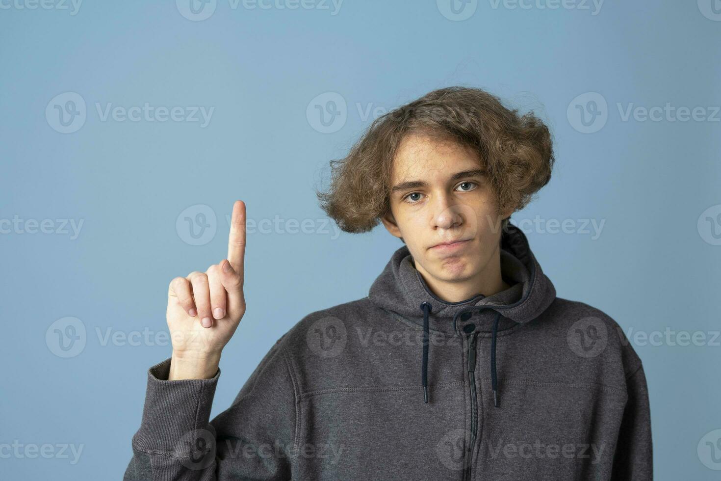 une adolescent dans une gris sweat à capuche avec ondulé cheveux points en haut avec le sien indice doigt sur une bleu Contexte photo