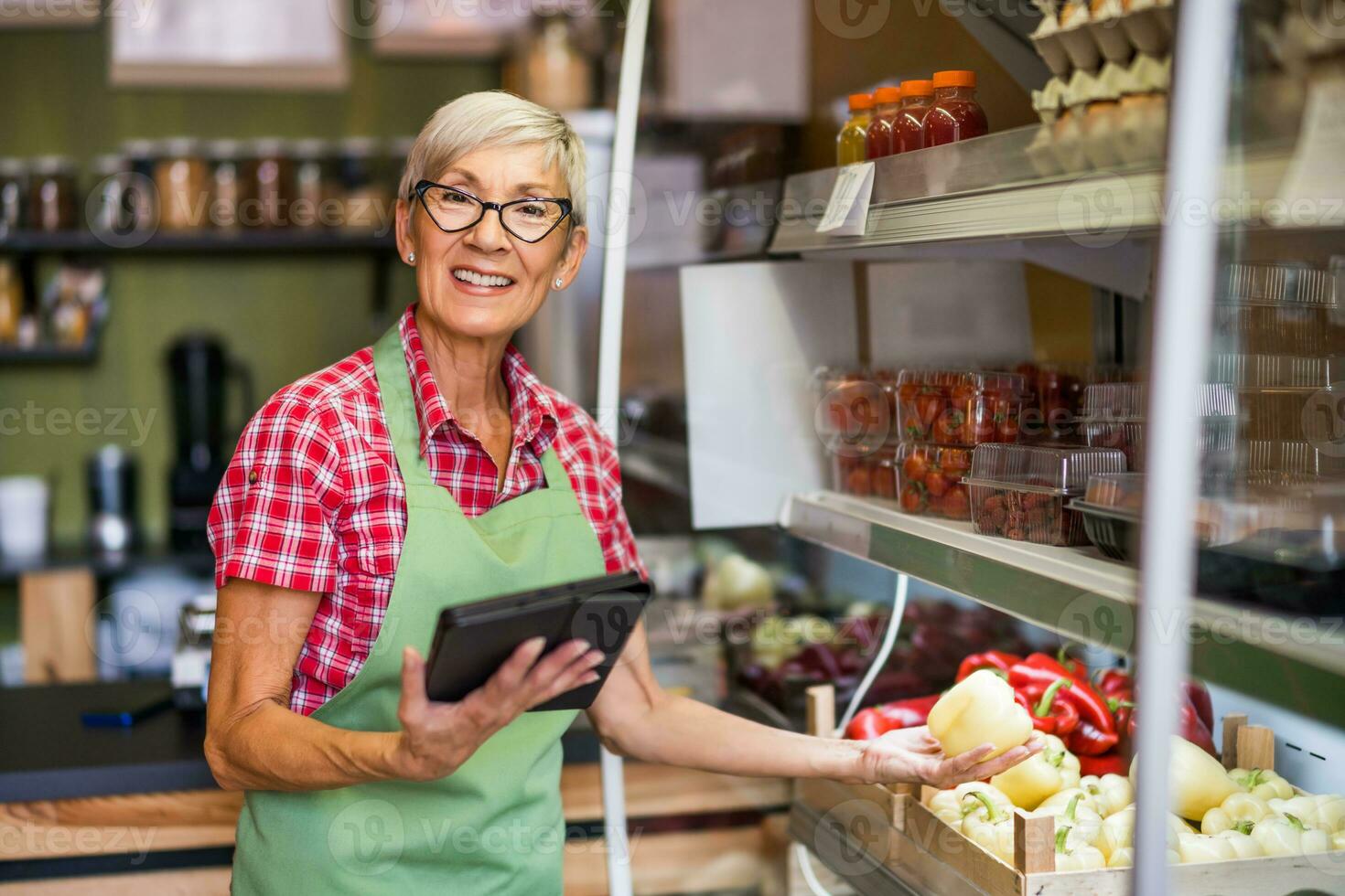 Sénior femme travaux dans des fruits et des légumes magasin. elle est en portant une tablette dispositif et Jaune poivrons.. photo